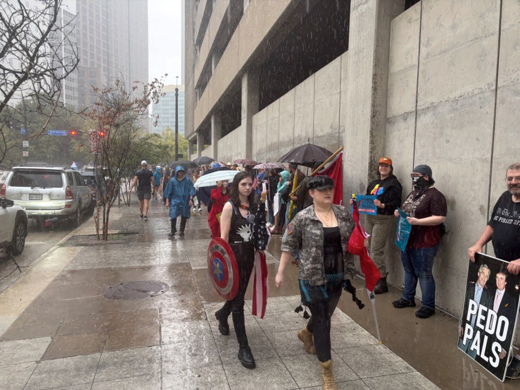 No Kings protesters in Dallas shelter under a parking garage during rain.