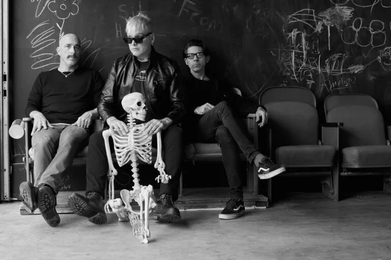 The band members of Alkaline Trio pose with a skeleton in a black and white press photo.